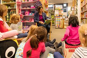 Eine Frau sitzt mit Kindern in einem Kreis und liest aus einem Buch vor.