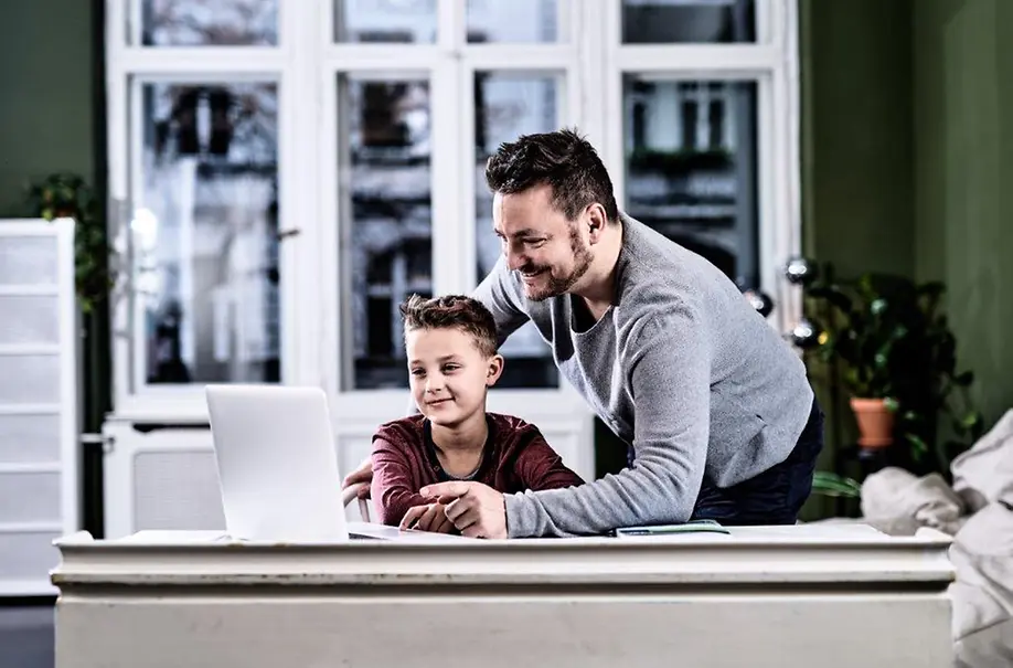 Ein Junge und ein Vater sitzen an einem Schreibtisch und schauen gemeinsam auf einen Laptop.
